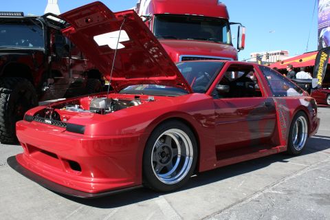 1998 Ford Crown Victoria - red widebody sport coupe with its hood up and polished chrome wheels sits on pavement in front of semi-trailers at an outdoor show.