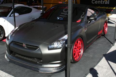 1998 Ford Crown Victoria - matte gunmetal Nissan GT-R with bright red wheels and carbon-fiber trim sits at an outdoor display booth, a white GT-R behind.