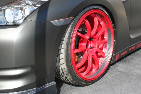 1998 Ford Crown Victoria - Close-up of a bright red multi-spoke wheel and low-profile tire on the front of a matte gunmetal Nissan GT-R with carbon trim.