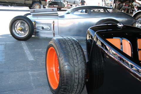 1998 Ford Crown Victoria - bare-metal lakester-style hot rod with wire wheels sits beside a black roadster with orange wheels and tan interior on pavement.