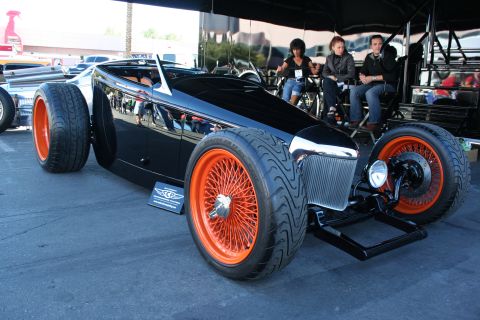 1998 Ford Crown Victoria - low black custom roadster hot rod with orange wire wheels sits under a canopy as seated spectators watch at an outdoor car show.
