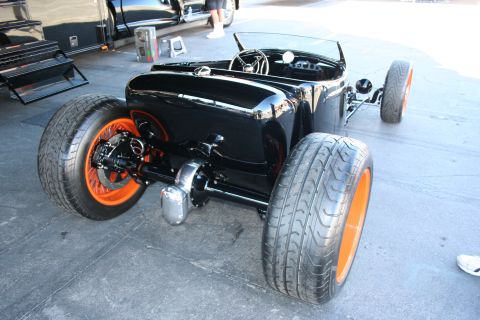 1998 Ford Crown Victoria - Rear view of a black open-cockpit hot rod roadster riding on staggered tires with bright orange wheels on a paved show lot.