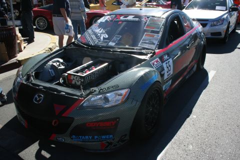 1998 Ford Crown Victoria - carbon-liveried Mazda RX-8 race car wearing number 55 and sponsor decals sits with its hood raised in a crowded show lane.