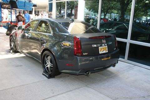 1998 Ford Crown Victoria - Rear three-quarter view of a gray Cadillac CTS-V sedan with KPE sponsor decals parked outside a glass-fronted building.
