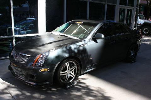 1998 Ford Crown Victoria - Front three-quarter view of a dark gray Cadillac CTS-V sedan on black wheels with KPE splitters beside a glass-fronted showroom.