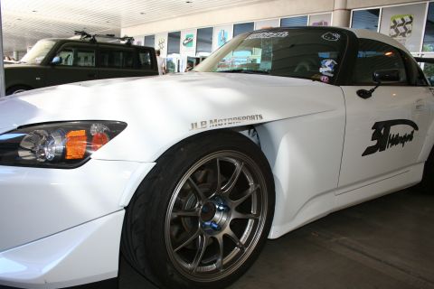 1998 Ford Crown Victoria - white Honda S2000 roadster with widened fenders, bronze multi-spoke wheels and JLB Motorsports graphics parked under a covered showroom ent…