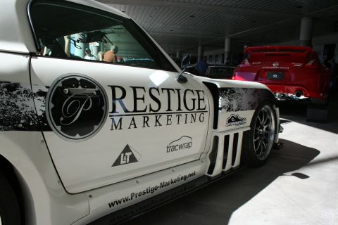 1998 Ford Crown Victoria - Close-up of a white widebody race car door wrapped in Prestige Marketing graphics, with a red coupe and multi-spoke wheel visible behind.