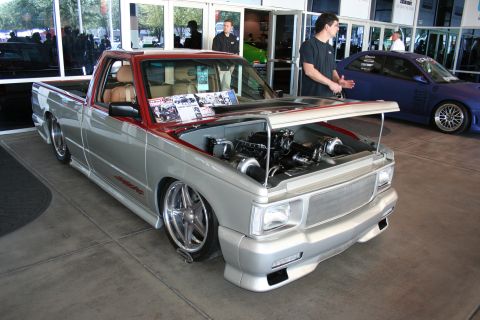 1998 Ford Crown Victoria - lowered silver pickup truck with red accents and a raised hood revealing a twin-turbocharged engine, displayed outside a glass storefront.