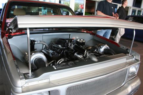 1998 Ford Crown Victoria - Close-up of a custom truck engine bay with twin large turbochargers and intricate plumbing beneath a raised hood, people standing behind.