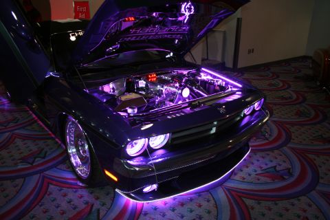 1998 Ford Crown Victoria - dark Dodge Challenger with raised hood and purple LED underglow and engine lighting, displayed on patterned convention carpet.