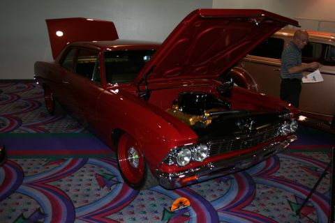 1998 Ford Crown Victoria - dark red classic American muscle coupe with both hood and trunk open displayed on patterned convention carpet, a man examining it.