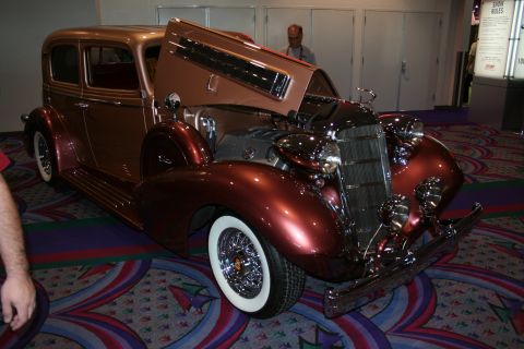 1998 Ford Crown Victoria - copper-brown two-tone 1930s sedan with wire wheels, wide whitewall tires and an open hood displayed on patterned convention carpet.