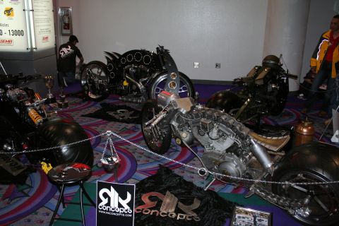 1998 Ford Crown Victoria - Several elaborate custom chopper motorcycles displayed behind a chain barrier on patterned carpet with RK Concepts signage.