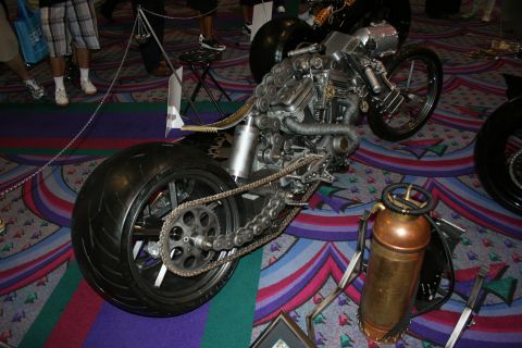 1998 Ford Crown Victoria - bare-metal custom chopper motorcycle with an exposed engine, fat rear tire and long drive chain displayed on patterned carpet.
