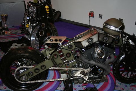 1998 Ford Crown Victoria - olive-green custom chopper with an exposed V-twin engine, bobber-style frame and chain drive displayed on patterned carpet indoors.