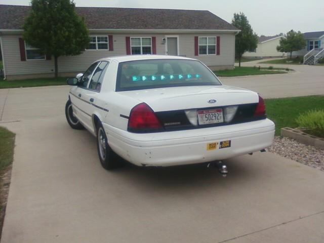 White Ford Crown Victoria sedan from rear, interior light bar glowing in rear window, parked in driveway