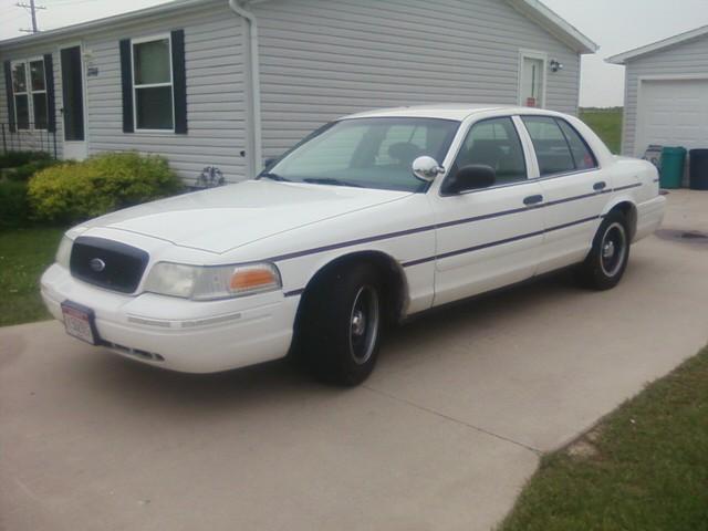 White Ford Crown Victoria sedan with black steel wheels and spotlight, parked on a driveway, front 3/4 view