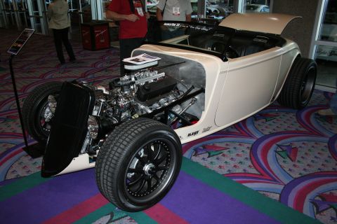 1998 Ford Crown Victoria - cream-colored 1930s-style hot rod roadster with an exposed chrome V8 engine, open wheels and black rims displayed indoors.