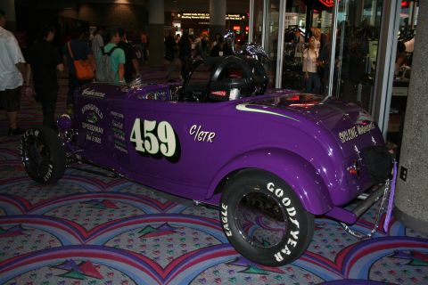 1998 Ford Crown Victoria - purple open-wheel hot rod race car marked 459 with Goodyear tires and lettering, parked on patterned carpet near a glass storefront.