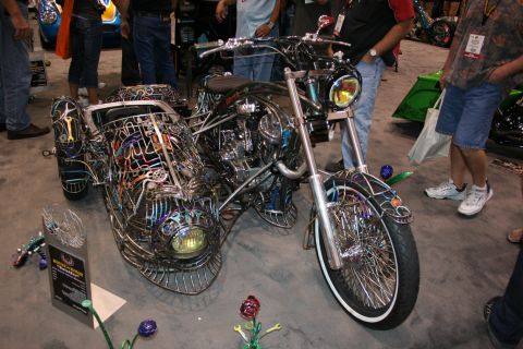 1998 Ford Crown Victoria - custom three-wheeled motorcycle built from intricate welded metal rods and multicolored tubing forming an open skeletal body, on a show flo…