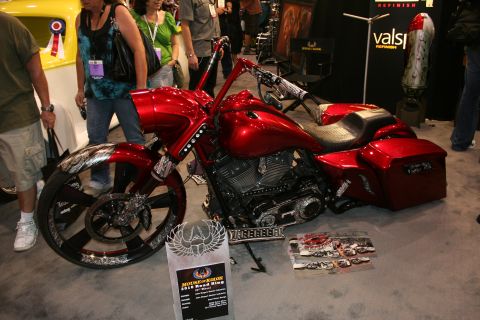 1998 Ford Crown Victoria - candy-red custom bagger motorcycle with a tall front wheel, stretched bags and a winged trophy placard on the floor at an indoor show.