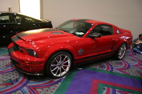 1998 Ford Crown Victoria - glossy red modern Mustang coupe with silver stripes, hood vents, a snake emblem and polished wheels parked on a patterned carpet indoors.