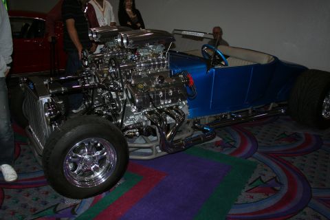 1998 Ford Crown Victoria - blue open T-bucket hot rod with exposed twin supercharged chrome engines and a white interior, parked on patterned carpet indoors.