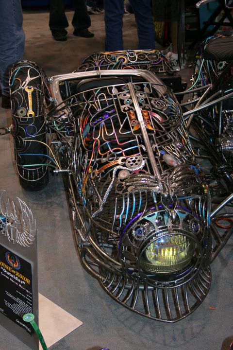 1998 Ford Crown Victoria - Close-up of an intricate welded-metal custom trike with exposed colorful tubing, a single glowing round headlight and a wing-shaped placard…