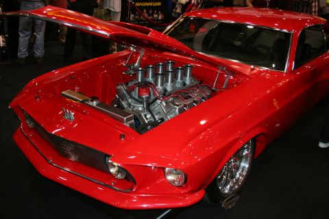 1998 Ford Crown Victoria - glossy red fastback muscle car displayed with its hood up, revealing a polished V8 topped with eight individual velocity stacks.