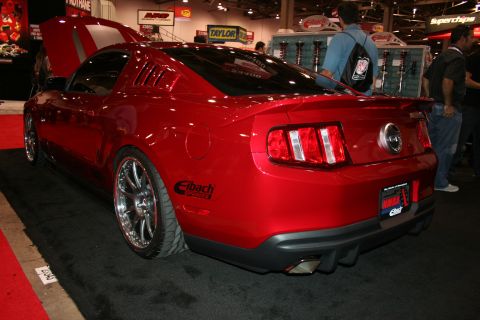 1998 Ford Crown Victoria - rear three-quarter view of a candy-red modern Mustang at an indoor expo, lowered on chrome wheels with an Eibach side decal.