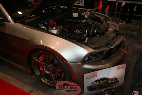 1998 Ford Crown Victoria - silver-grey Mustang sits with its hood open at an indoor show beside an Air Lift suspension display board.