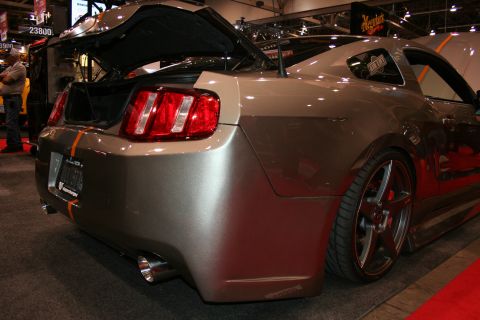 1998 Ford Crown Victoria - Rear three-quarter view of a grey modern Mustang with an orange accent stripe and open decklid, lowered on bronze wheels at a show.