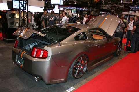 1998 Ford Crown Victoria - grey modern Mustang with orange racing stripes sits with its hood and trunk open on a red carpet at a crowded indoor expo.