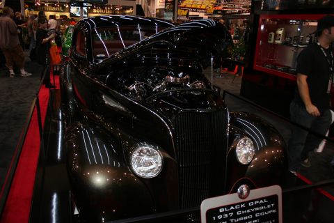 1998 Ford Crown Victoria - glossy black 1937 Chevrolet coupe shown front-on at an indoor expo, with a placard reading Black Opal 1937 Chevrolet.