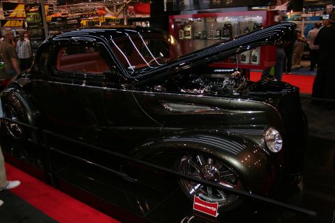 1998 Ford Crown Victoria - glossy black chopped hot rod coupe shown in side profile with its hood open, lowered on chrome wheels on a red carpet at a show.