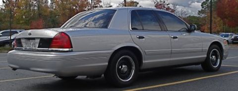 2000 Ford Crown Victoria - A silver 2000 Ford Crown Victoria sedan is parked in a lot, viewed from the rear three-quarter angle with autumn foliage in the background.