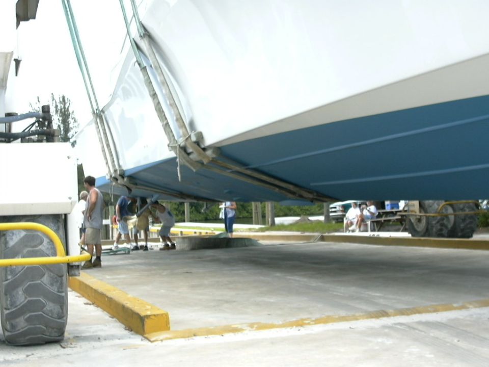 White boat with blue hull bottom hanging in travel lift slings at a marina, people underneath on concrete lot