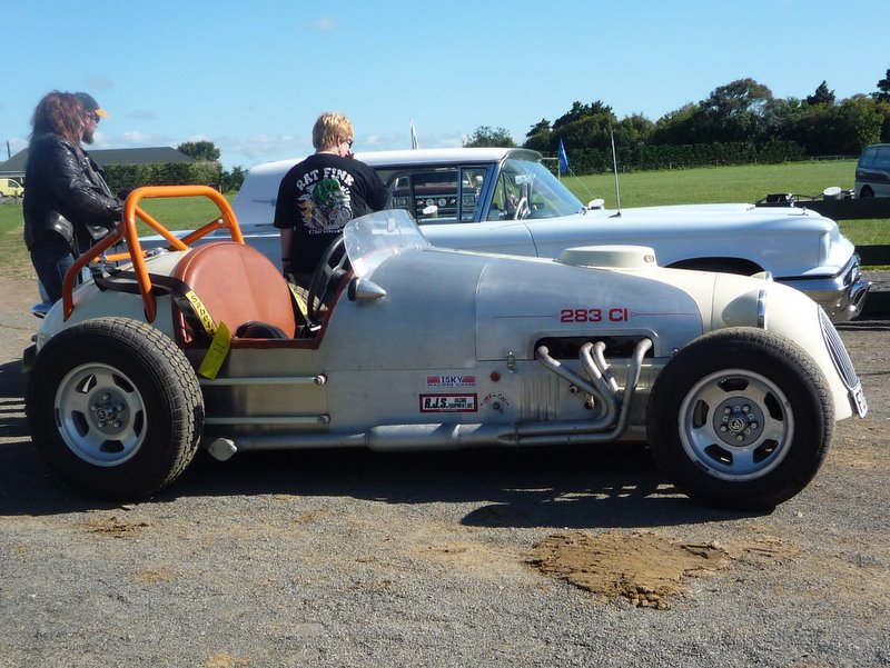 Side view of a silver open-wheel race car with a roll cage and a white car behind it