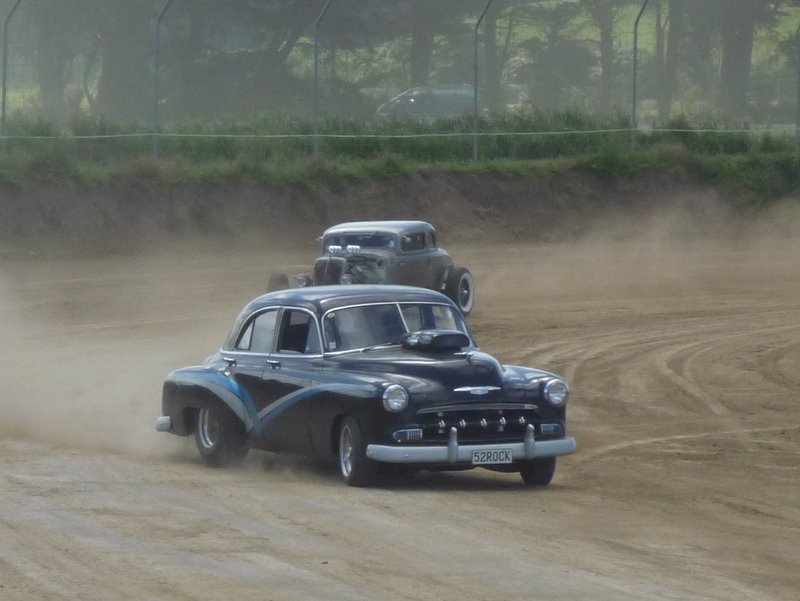 Black hot rod with blue accents on a dirt track, kicking up dust. License plate reads 52ROCK