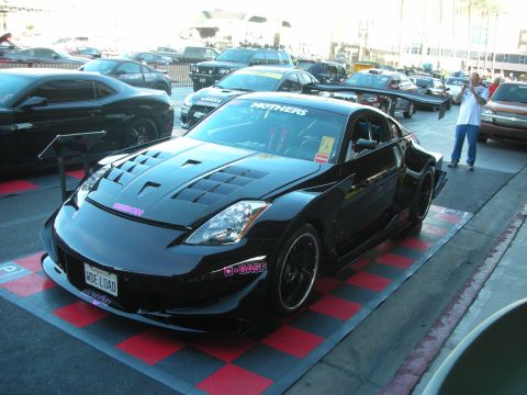 1998 Ford Crown Victoria - black Nissan 350Z with a vented hood, body kit, and tall rear wing sits on a red checkered display pad in an outdoor show.