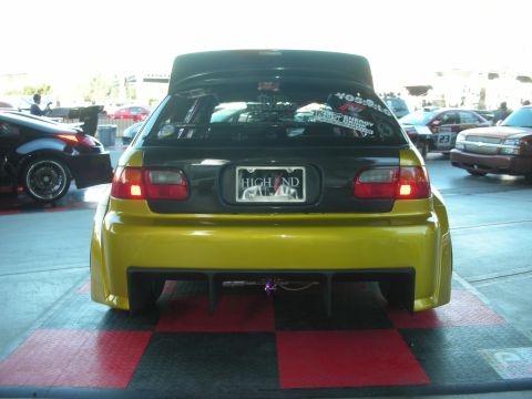 1998 Ford Crown Victoria - rear of a yellow tuner hatchback with a large wing, rear diffuser, and a HIGH END license plate, parked on a checkered show pad.