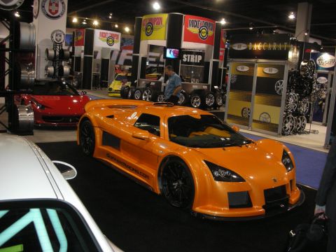 1998 Ford Crown Victoria - orange Gumpert Apollo supercar parked on a show floor near a red Ferrari and wheel-brand booths.