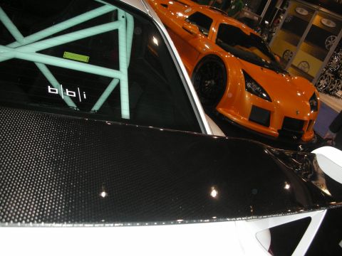 1998 Ford Crown Victoria - Close view of a carbon-fiber rear deck and roll bar marked bbi, with an orange supercar visible through the glass at an indoor show.