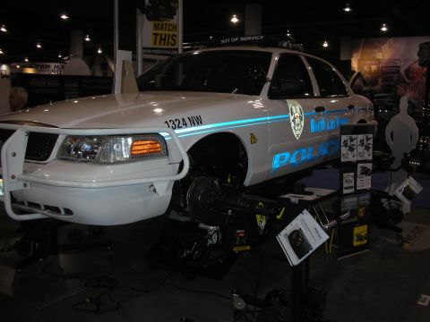 1998 Ford Crown Victoria - white Crown Victoria-style police interceptor displayed on an alignment rack with front wheels removed at an indoor equipment booth.