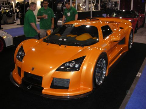 1998 Ford Crown Victoria - orange Gumpert Apollo supercar with quad projector headlights and a tan cockpit on a show stand as green-shirted staff stand nearby.