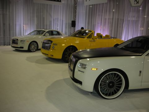 1998 Ford Crown Victoria - trio of customized Rolls-Royce cars on a draped showroom floor: a white sedan, a yellow convertible and a white car with black roof on larg…