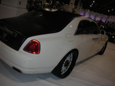 1998 Ford Crown Victoria - Rear three-quarter view of a white Rolls-Royce Ghost with a black roof, dual exhaust and black multi-spoke wheels on a show floor.
