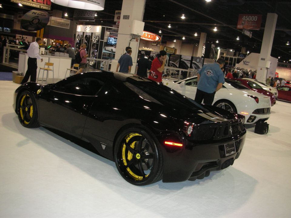 Black Ferrari 458 with yellow-accent wheels at busy indoor show floor