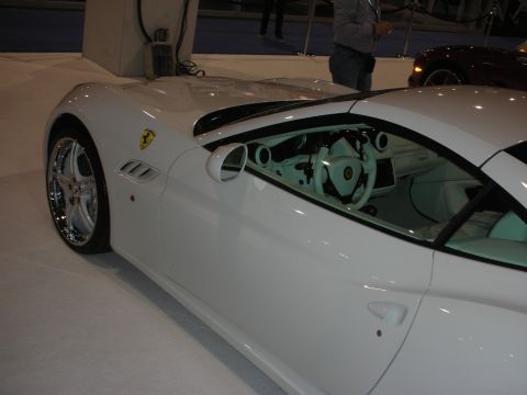 1998 Ford Crown Victoria - Side view of a white Ferrari coupe with a pale interior, chrome multi-spoke wheel and yellow prancing-horse fender badge at an indoor show.
