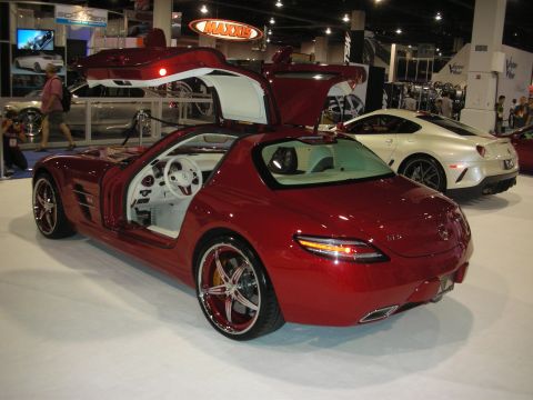 1998 Ford Crown Victoria - red Mercedes-Benz SLS AMG with one gullwing door raised, white interior and chrome wheels, displayed beside a white coupe at an indoor show.